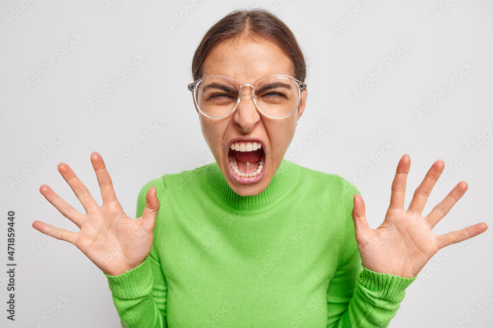 Hysterical mad European woman feels crazy keeps palms raised screams loudly from anger wears green jumper and big transparent glasses isolated over white background. Negative emotions concept Hysterical mad European woman feels crazy keeps palms raised screams loudly from anger wears green jumper and big transparent glasses isolated over white background. Negative emotions concept