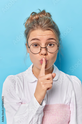 Image of pretty mysterious woman presses index finger over lips makes hush gesture shows taboo gesture asks to be quiet dressed in neat clothes round spectacles isolated over blue background