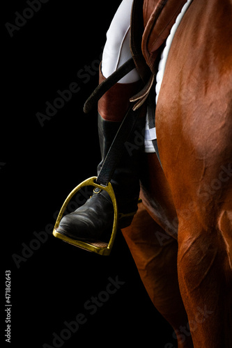 A rider's foot on brown horse looking forward closeup. A woman's horse riding booted foot standing in a gold stirrup of horse saddle isolated on black background. photo