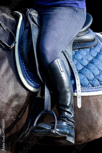 A rider's foot on black horse closeup isolated on black background. A woman's booted foot standing in a black stirrup of horse saddle.