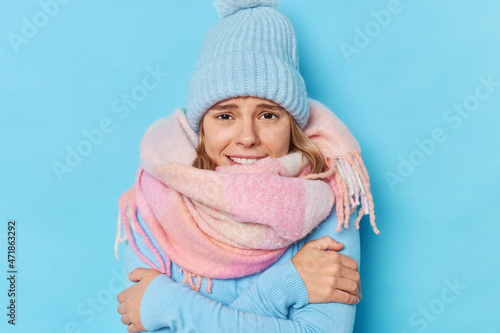 Brr how freezing there. Young woman cuddles and feels cold asks to turn on heater wears knitted hat warm scarf around neck walks during frozing winter day shivers isolated over blue background
