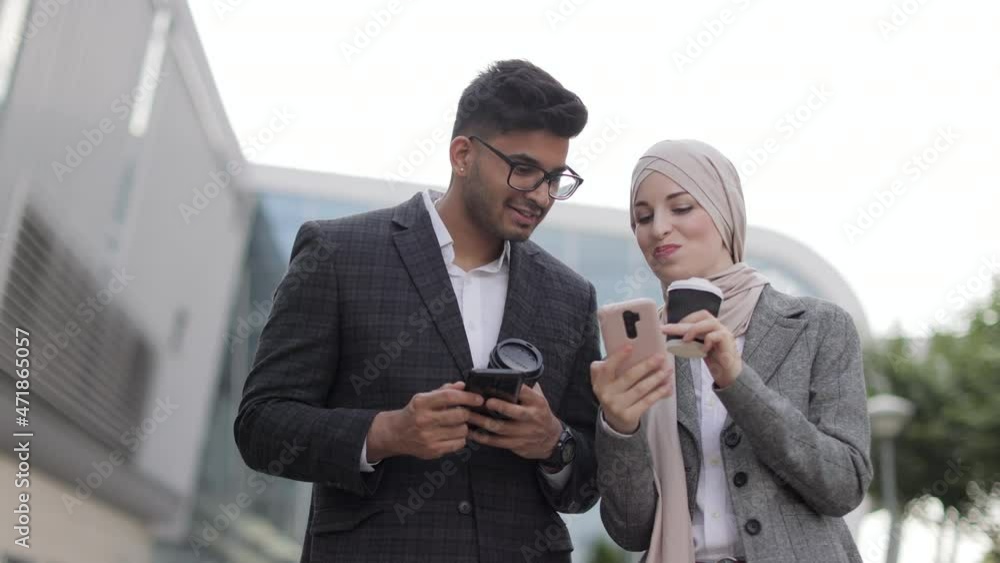 Arabian business couple having fun and talking together walking on the street during coffee break. Woman in hijab showing her smartphone screen to her colleague, sharing idea or news Arabian business couple having fun and talking together walking on the street during coffee break. Woman in hijab showing her smartphone screen to her colleague, sharing idea or news