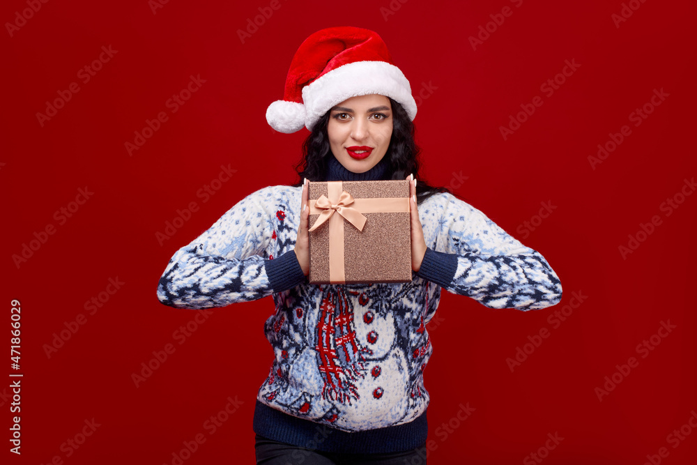Fototapeta premium A cute young girl in a Christmas cap on a red background holds a gift box in front of her. Space for text.