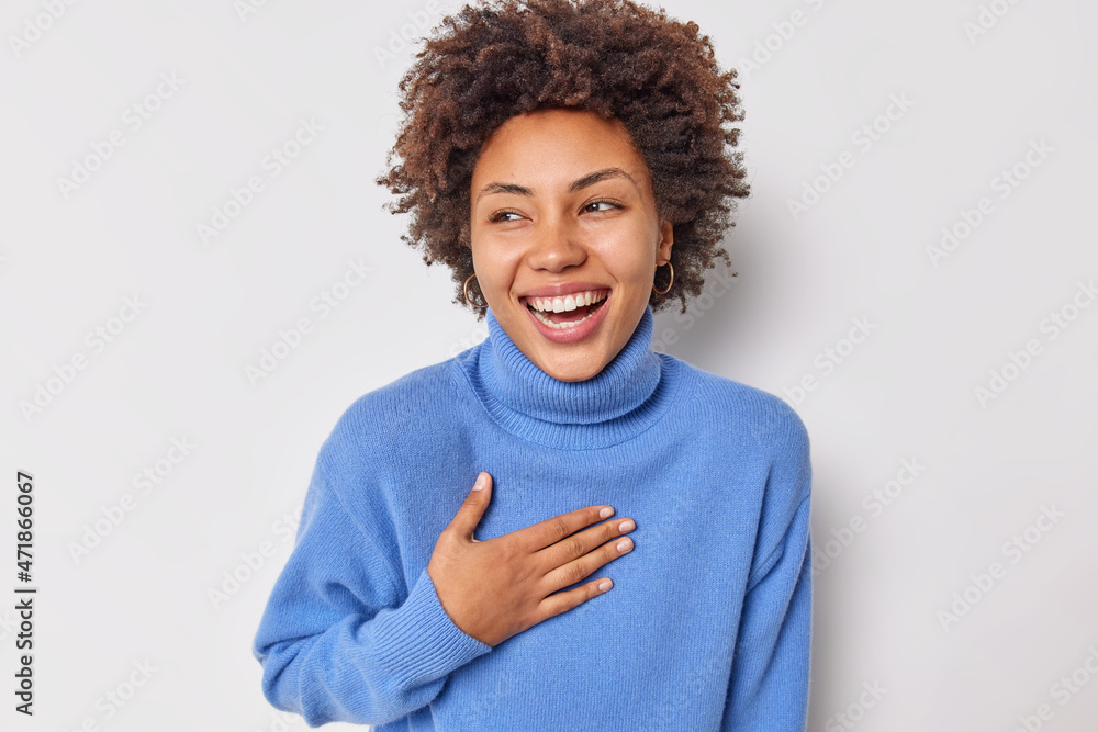 Portrait of cheerful curly haired woman smiles broadly keeps hand on chest cannot stop laughing at funny joke wears casual blue jumper isolated over white background. Positive human emotions concept Portrait of cheerful curly haired woman smiles broadly keeps hand on chest cannot stop laughing at funny joke wears casual blue jumper isolated over white background. Positive human emotions concept