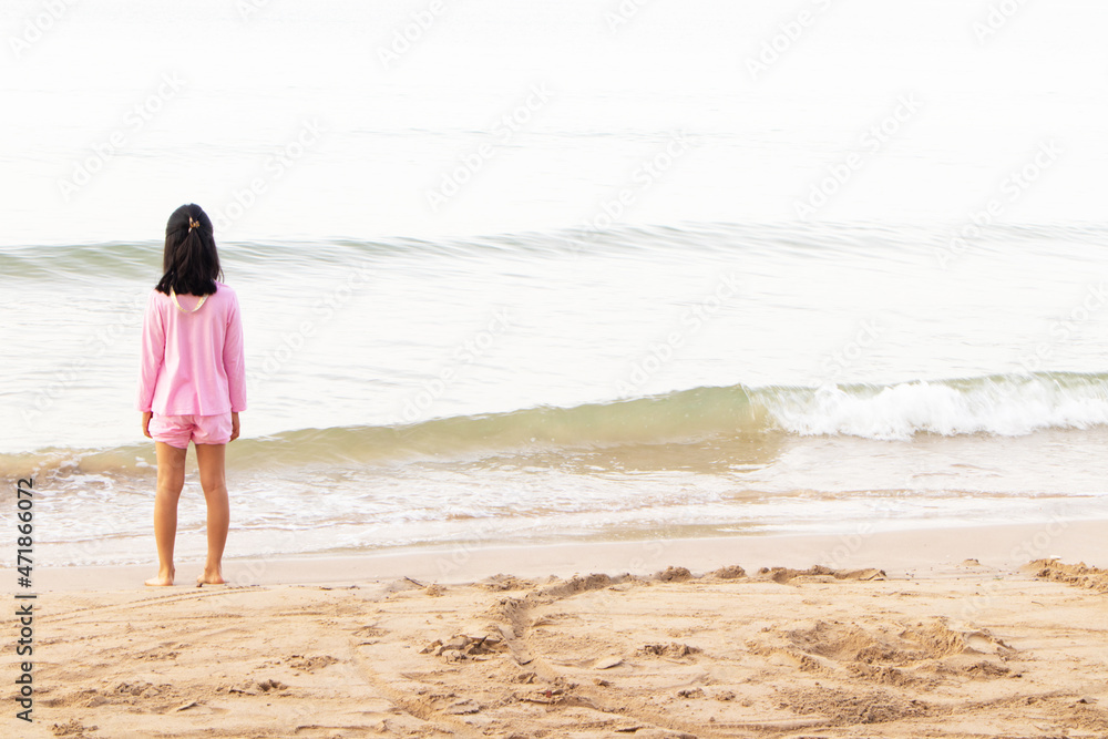 Cute toddler Asian girl standing alone on brown beach at bright sunlight. Child standing on beach near sea and looking on waves.  Solitude concept. Retreat leisure on summer family vacation with kids.