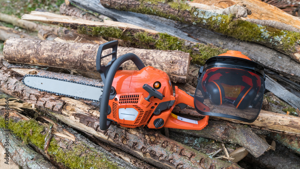 Portable power chain saw and safety helmet on heap of mossy wood logs. Orange chainsaw and hard hat with face guard and hearing protection. Occupational safety and health at work with sawing machine. Portable power chain saw and safety helmet on heap of mossy wood logs. Orange chainsaw and hard hat with face guard and hearing protection. Occupational safety and health at work with sawing machine.