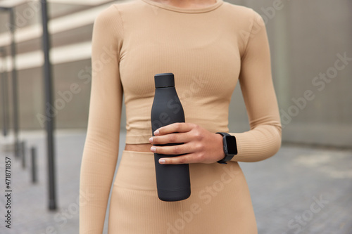 Cropped shot of unrecognizable woman dressed in active wear holds bottle with fresh water leads active lifestyle takes break after doing sport exersices trains outdoors. People and physcial activity