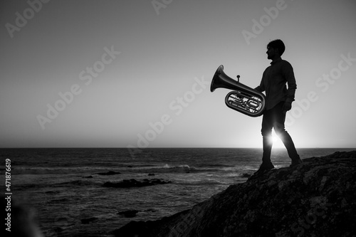 Silhouette of a man with a trumpet by the ocean during sunset. Black and white photo.
