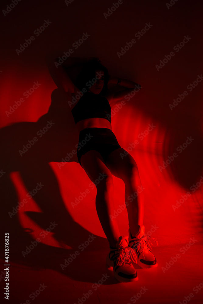 Photoshoot of a young brunette in a photo studio. Beautiful slender woman in a black top and shorts in red light.