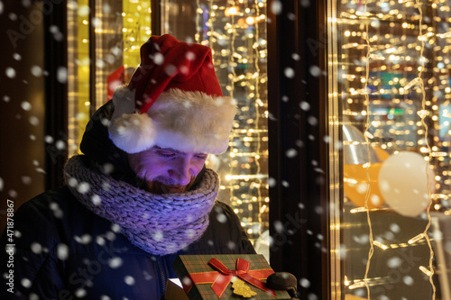 Fototapeta Naklejka Na Ścianę i Meble -  Man in Santas hat with gifts box near illuminated cafe window.