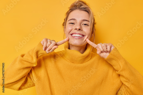 Portrait of cheerful young woman smiles broadly indicates index fingers at face wears casual jumper expresses positive emotions isolated over vivid yellow background. Look at my brilliant smile