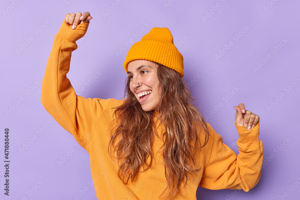 Positive carefree woman full of cool energy has fun celebrates win or victory makes triumph dance smiles joyfully wears casual orange jumper and hat isolated over purple background. People and fun