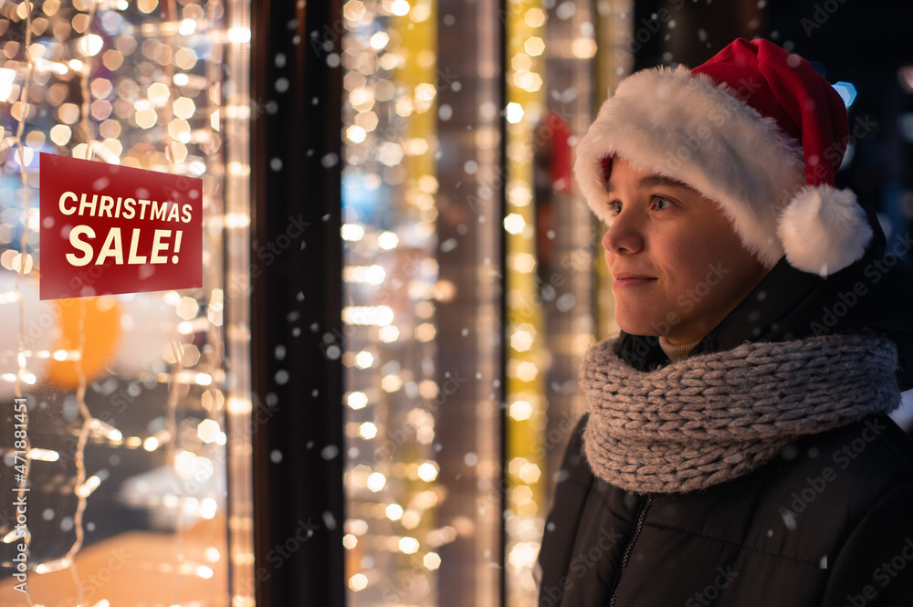 Boy in Santas hat looking and dreaming in illuminated shop window. Christmas holidays sales concept