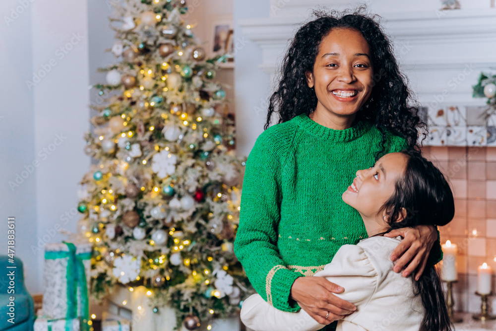 African American curly woman hugs brunette long-haired daughter and smiles against decorated Christmas tree in living room