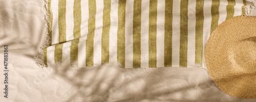 Striped linen beach towel with fringes and straw hat on sandy beach with shadows from palm tree. Relaxation and tropical summer holidays concept