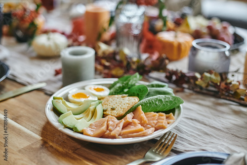 Plate with sliced fresh avocado, leaves of spinach, halved boiled egg, cookie and chopped fish standing on served wooden table