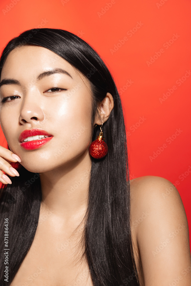 Fototapeta premium Vertical portrait of sexy, sensual asian woman wears christmas earrings, xmas tree decor in ear, gazing sensual at camera, red background