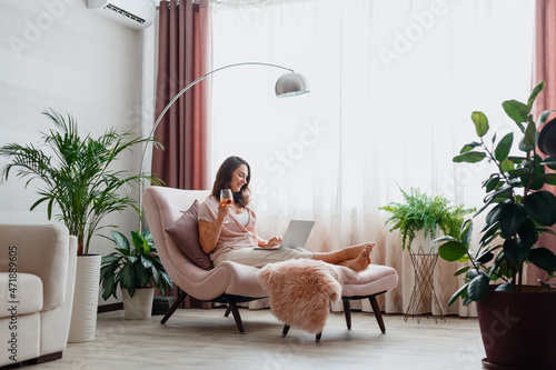Young beautiful woman drinking rose wine and using laptop while sitting on comfortable pink armchair in home interior near window. Relax woman watching movie on laptop or make online purchases.