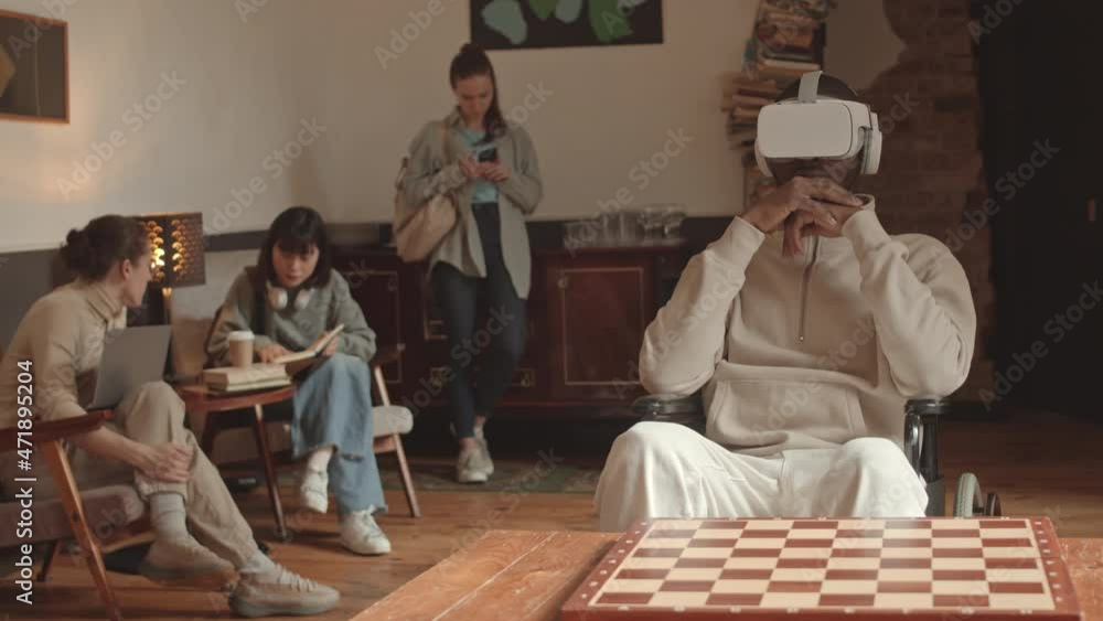 Medium slowmo shot of African-American male university student playing chess in VR headset while his classmates talking and studying together in background at modern campus Medium slowmo shot of African-American male university student playing chess in VR headset while his classmates talking and studying together in background at modern campus