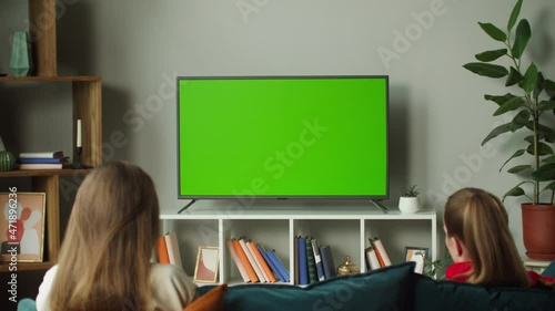 Women watching television with chroma green screen. Girlfriends switching channels on tv with remote controller, sitting on sofa together in living room. Spare time at home, relaxing. photo