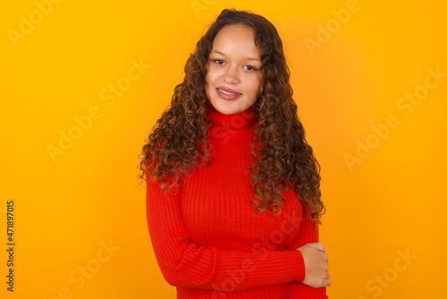 Teenager girl with curly hair wearing red sweater over yellow background laughing.