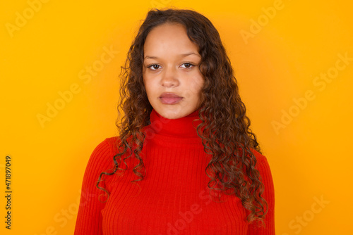 Joyful Teenager girl with curly hair wearing red sweater over yellow background looking to the camera, thinking about something. Both arms down, neutral facial expression. © Jihan