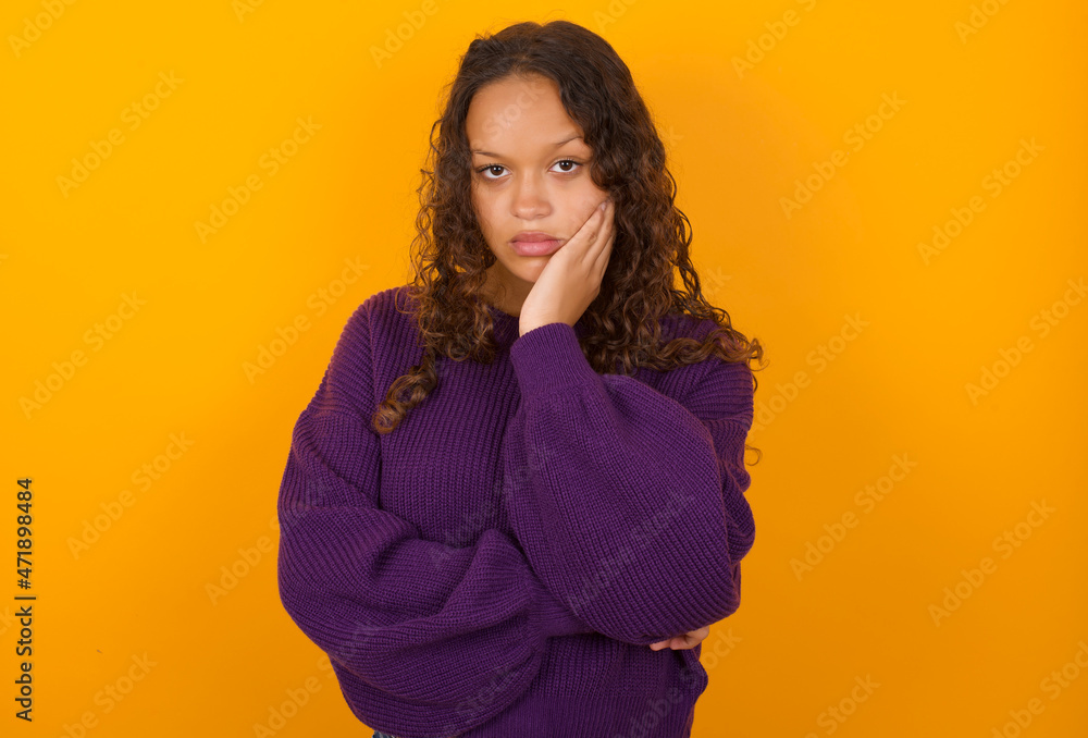 Very bored teenager girl wearing purple knitted sweater against yellow background holding hand on cheek while support it with another crossed hand, looking tired and sick. Very bored teenager girl wearing purple knitted sweater against yellow background holding hand on cheek while support it with another crossed hand, looking tired and sick.