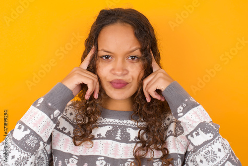 Serious concentrated teenager girl wearing winter knitted sweater against yellow wall keeps fingers on temples, tries to ease tension, gather with thoughts and remember important information for exam