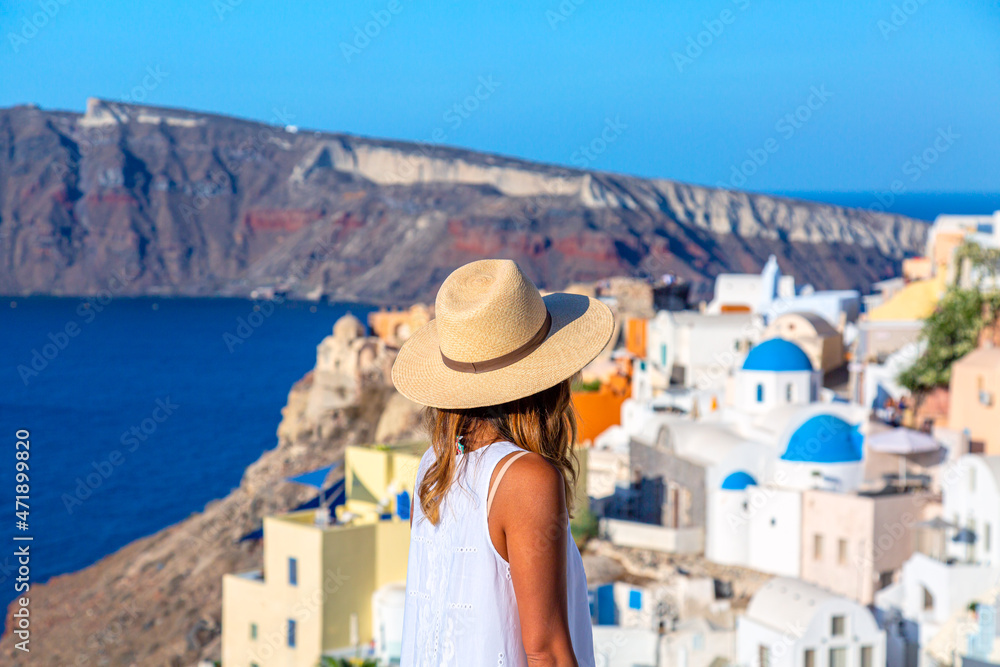 Beautiful tourist girl in straw hat against Oia village with famous white houses and blue churches on Santorini island, Aegean sea, Greece. Travel concept