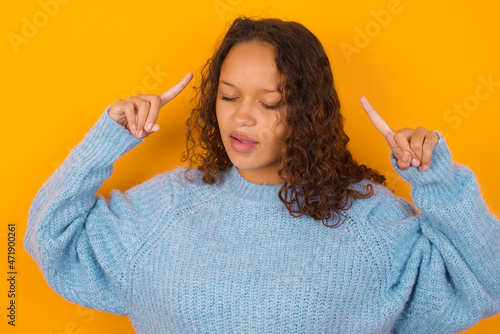 Photo of crazy Teenager girl wearing blue sweater over yellow background screaming and pointing with fingers at hair closed eyes
