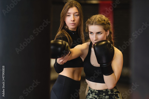 The blonde young woman, under the guidance of a trainer, performs blows on a punching bag. Women's fitness boxing workout in the gym.  © Volha