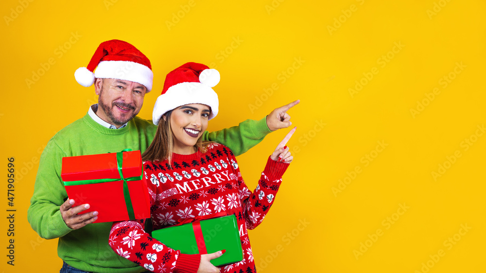 Obraz premium latin father and daughter in red sweater Christmas santa hat holding christmas presents pointing copy space on yellow background in Mexico Latin America