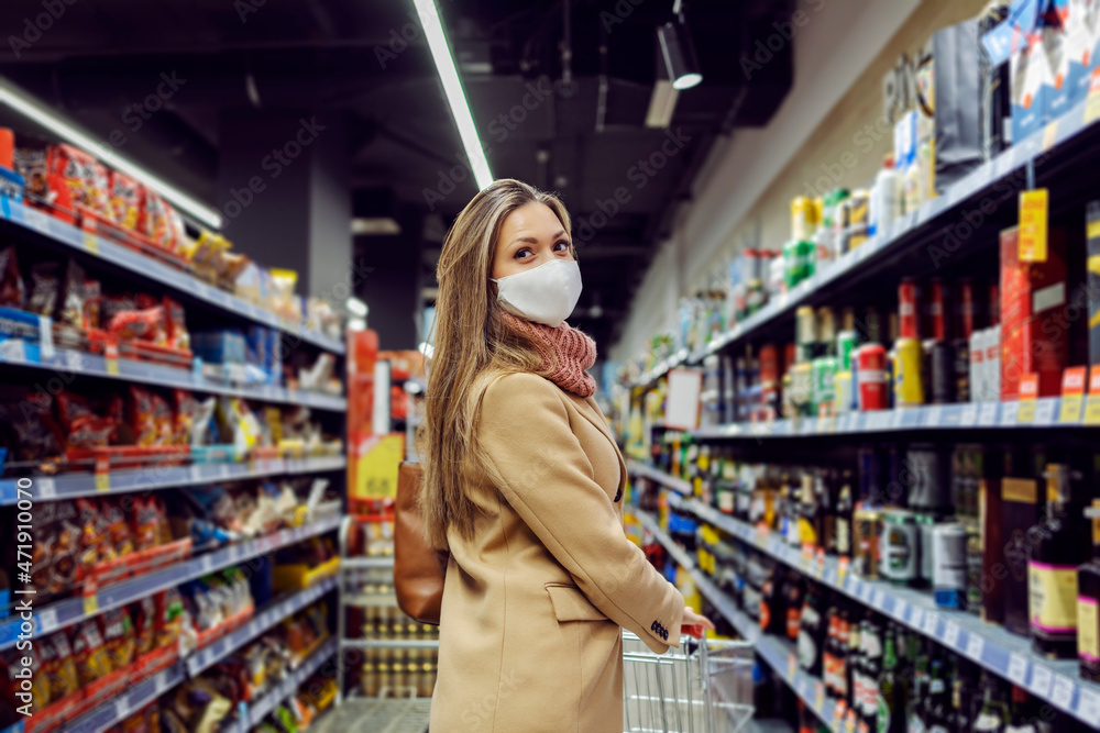 A woman with a face mask walks into the store and pushes the cart. The woman is going to buy some groceries and stuff for her home. Woman in the supermarket.
