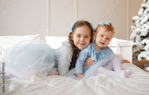 little sisters in blue holiday dresses are sitting on a bed in a room near a Christmas tree