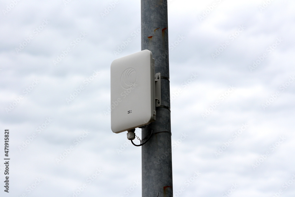 Modern new wireless communication device in white plastic case tied with plastic ties to old metal utility pole with rusted patches on dark gloomy cloudy sky background Modern new wireless communication device in white plastic case tied with plastic ties to old metal utility pole with rusted patches on dark gloomy cloudy sky background