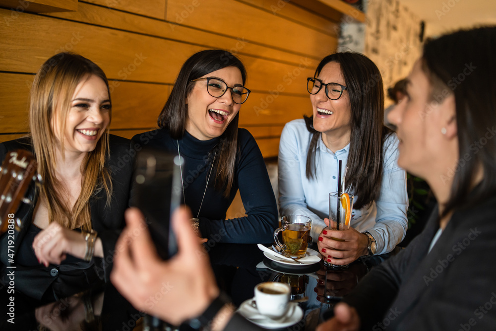 Front view of group of adult caucasian women young female holding mobile phone while sitting at cafe or restaurant with her millennial friends making a video call on smartphone or taking selfie photo