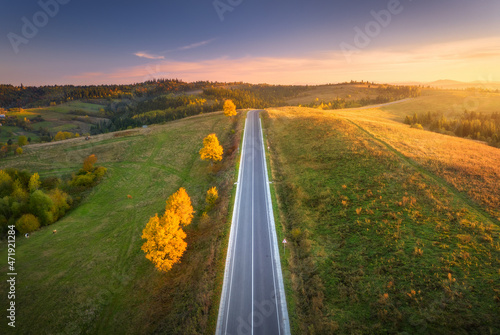 Aerial view of mountain road in green hills at sunset in autumn. Top view from drone of road. Beautiful landscape with roadway, orange trees, green meadows, blue sky, golden sunlight in fall. Travel