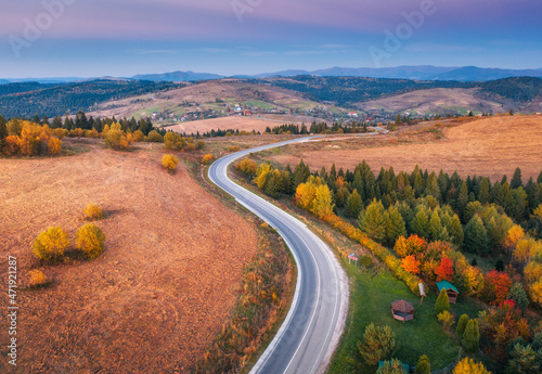 Aerial view of mountain road in forest at sunset in autumn. Top view from drone of road in woods at dusk. Beautiful landscape with roadway in hills, trees, meadows, fields, violet sky in fall. Travel