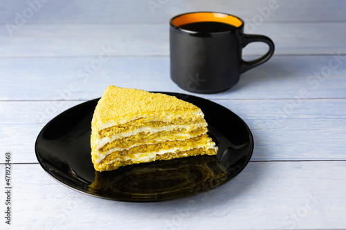 Piece of puff honey cake on black plate and cup nearby on blue plank surface photo