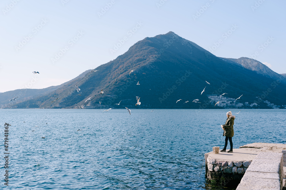 Girl in a jacket stands on the pier and feeds the seagulls against the backdrop of the mountains