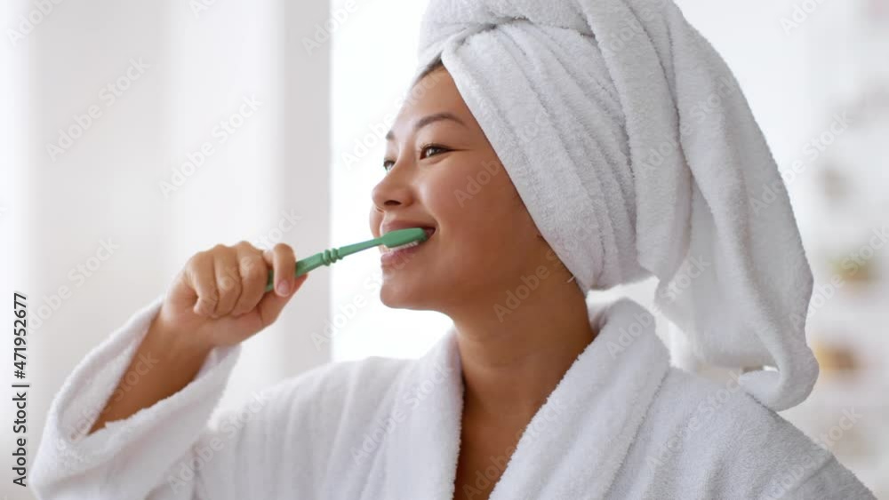 Morning routine. Young cheerful asian woman wearing bathrobe with towel on head brushing her teeth at bathroom Morning routine. Young cheerful asian woman wearing bathrobe with towel on head brushing her teeth at bathroom