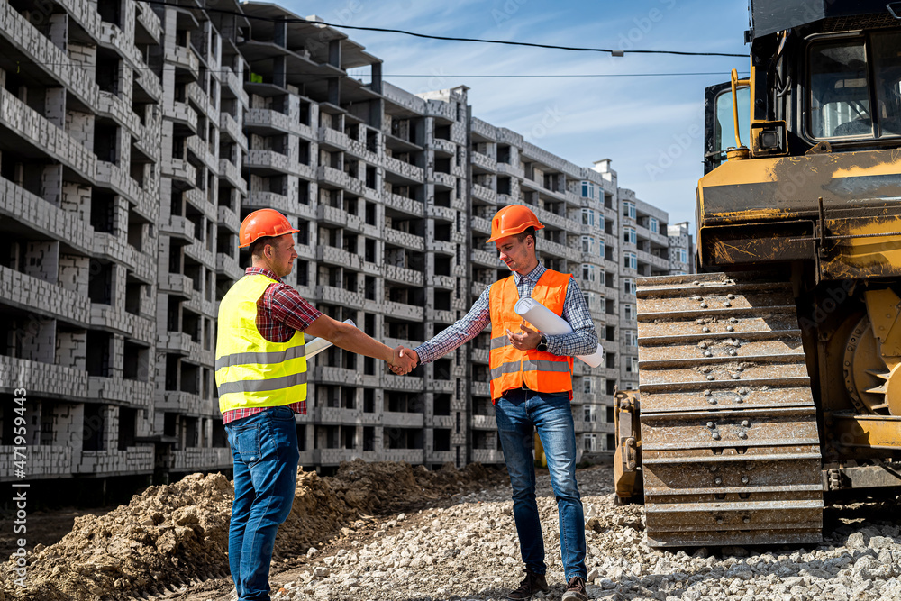 two good workers in uniform shake hands near the grader and the new building