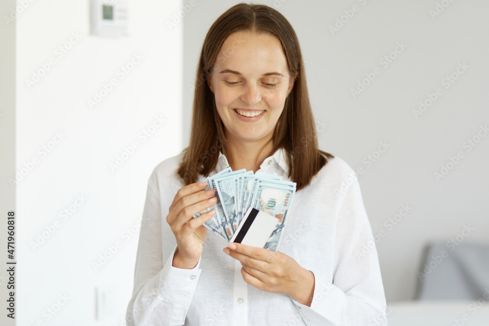 Portrait of smiling happy woman wearing white shirt standing indoor in light room, holding credit card and dollar banknotes, counting money and expressing positive emotions. Portrait of smiling happy woman wearing white shirt standing indoor in light room, holding credit card and dollar banknotes, counting money and expressing positive emotions.