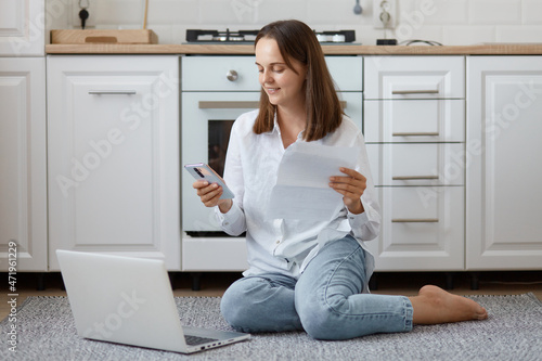 Free Download Indoor shot of smiling positive young adult female wearing white shirt and jeans, holding paper sheet and phone, working on computer, posing on floor in kitchen. - GFXLoad