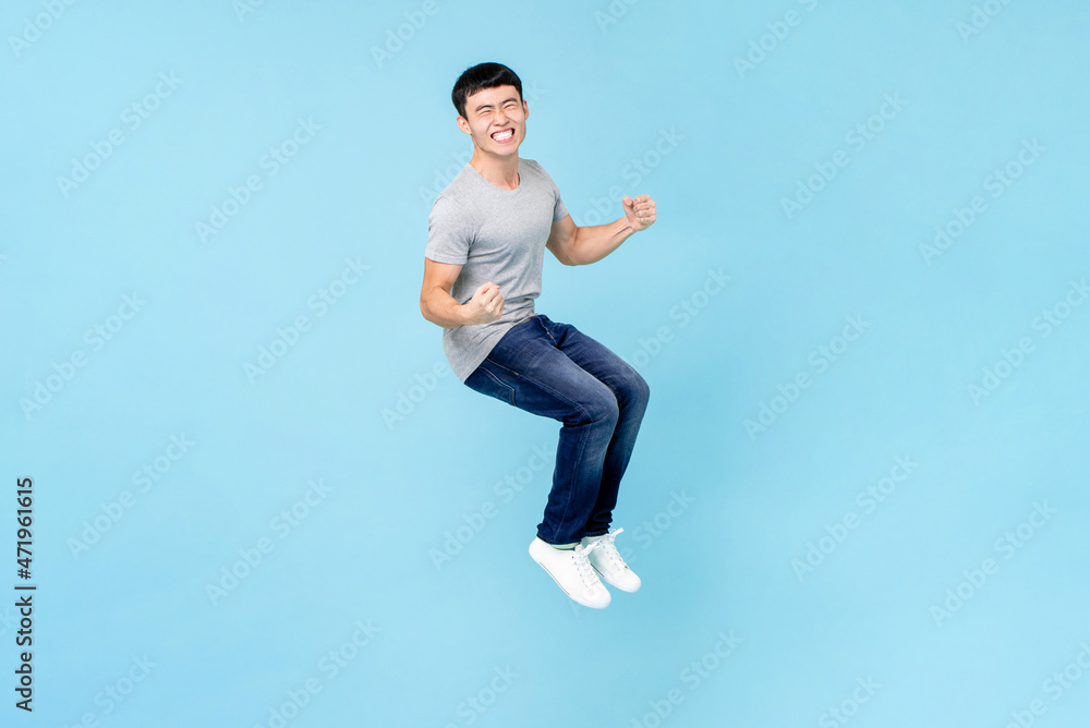 Portrait of smiling young happy Asian man clenching fists and jumping on isolated light blue studio background