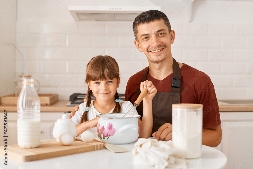 Indoor shot of smiling father and cute daughter posing in kitchen while sitting at table with products for making homemade pastry, family looking at camera with positive facial expression. Indoor shot of smiling father and cute daughter posing in kitchen while sitting at table with products for making homemade pastry, family looking at camera with positive facial expression.
