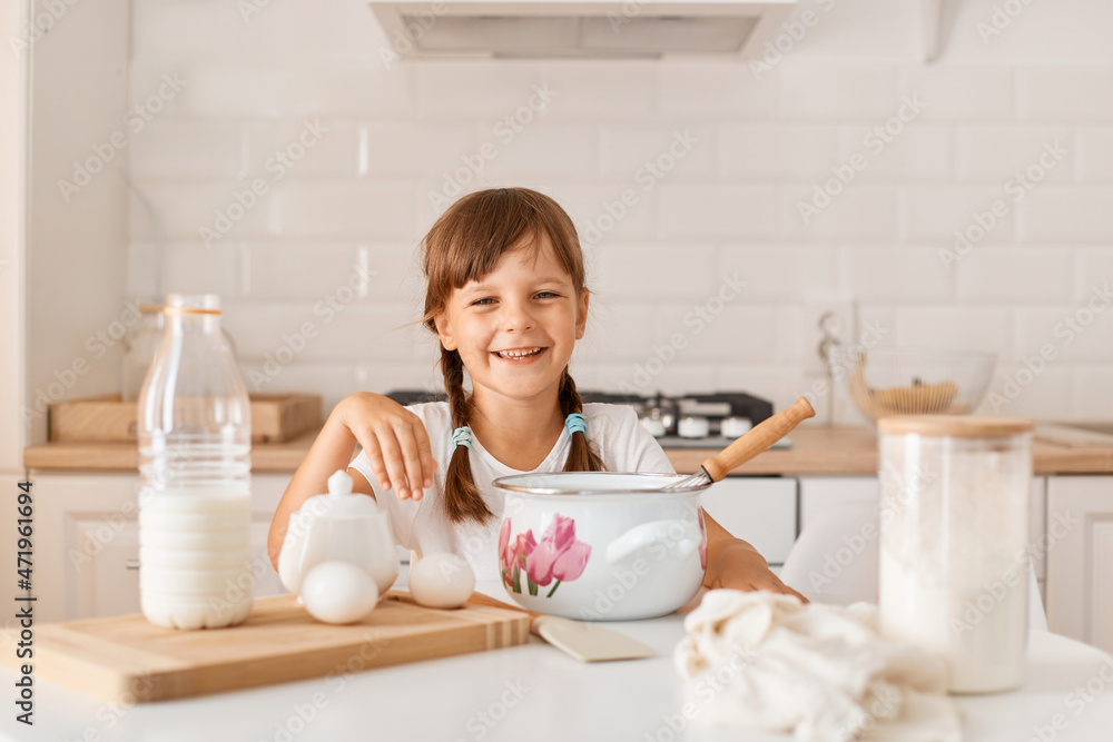 Portrait of smiling happy little girl with dark hair and ponytails sitting at table in kitchen and looking at camera, mixing eggs for pie with corolla in pot. Portrait of smiling happy little girl with dark hair and ponytails sitting at table in kitchen and looking at camera, mixing eggs for pie with corolla in pot.