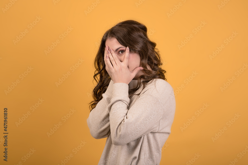 Caucasian beautiful young brunette girl in a casual jumper looking through her fingers, covering her face with her hands, isolated on an orange studio background.