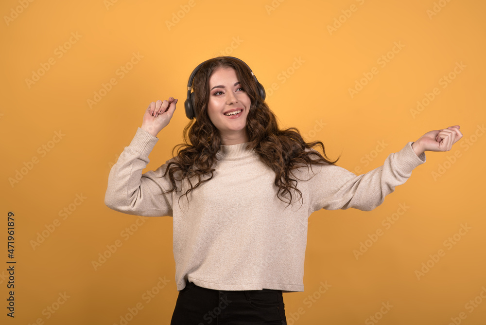 A young, attractive and happy Caucasian brunette girl with wavy hair listening to music on headphones and dancing on an orange studio background. A young, attractive and happy Caucasian brunette girl with wavy hair listening to music on headphones and dancing on an orange studio background.