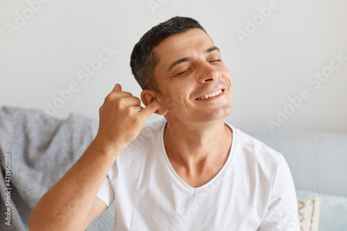Horizontal shot of smiling Caucasian brunette man wearing white t shirt, posing light room while sitting on gray couch and rubbing his ear, having satisfied facial expression.